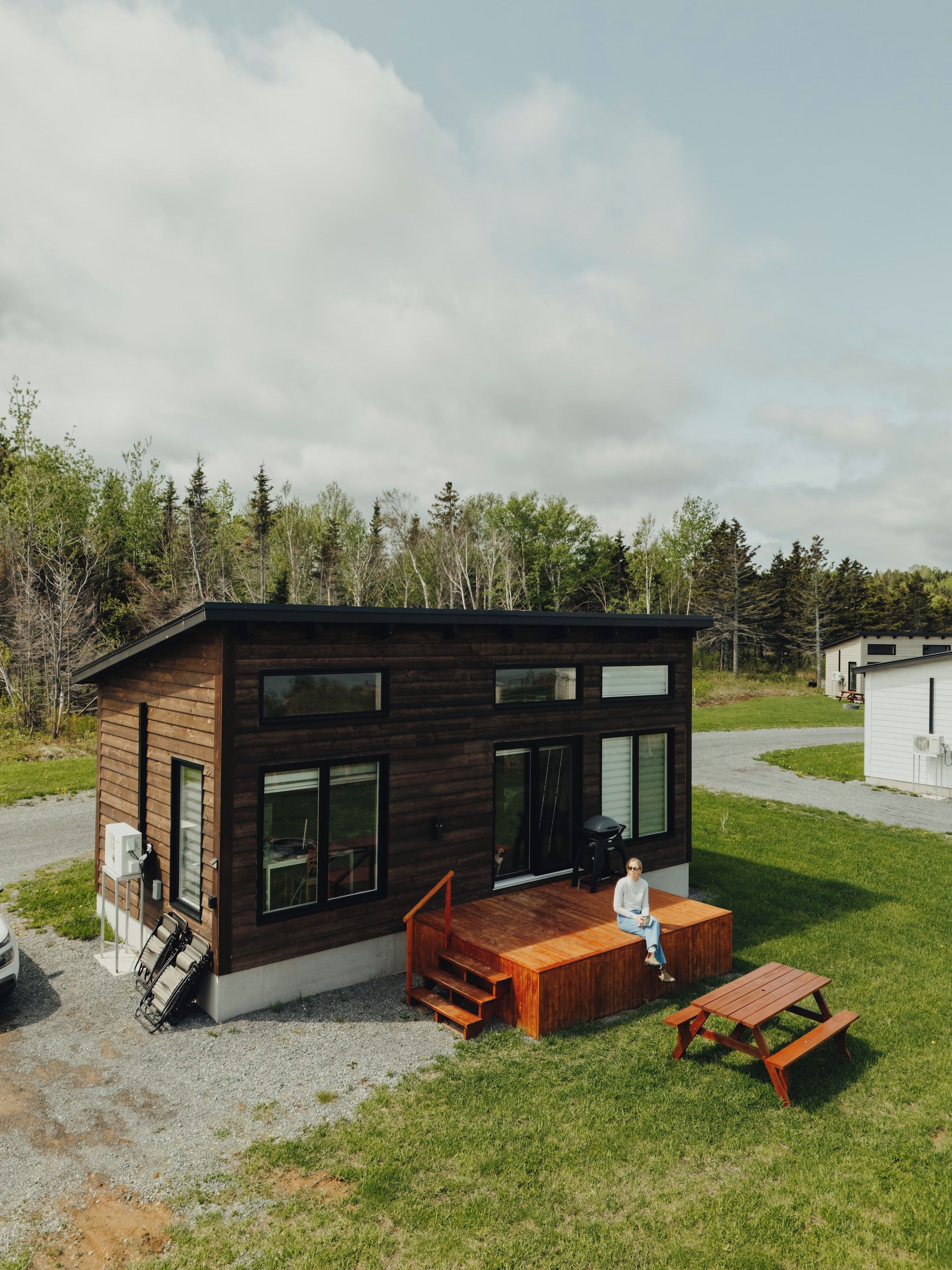 A modern wooden cabin with a deck, a small outdoor seating area, and a gravel driveway.