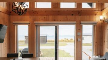 A spacious room with a high ceiling, wooden beams, a dining area with a table and chairs, a sofa, and large windows offering a view of the outdoors.