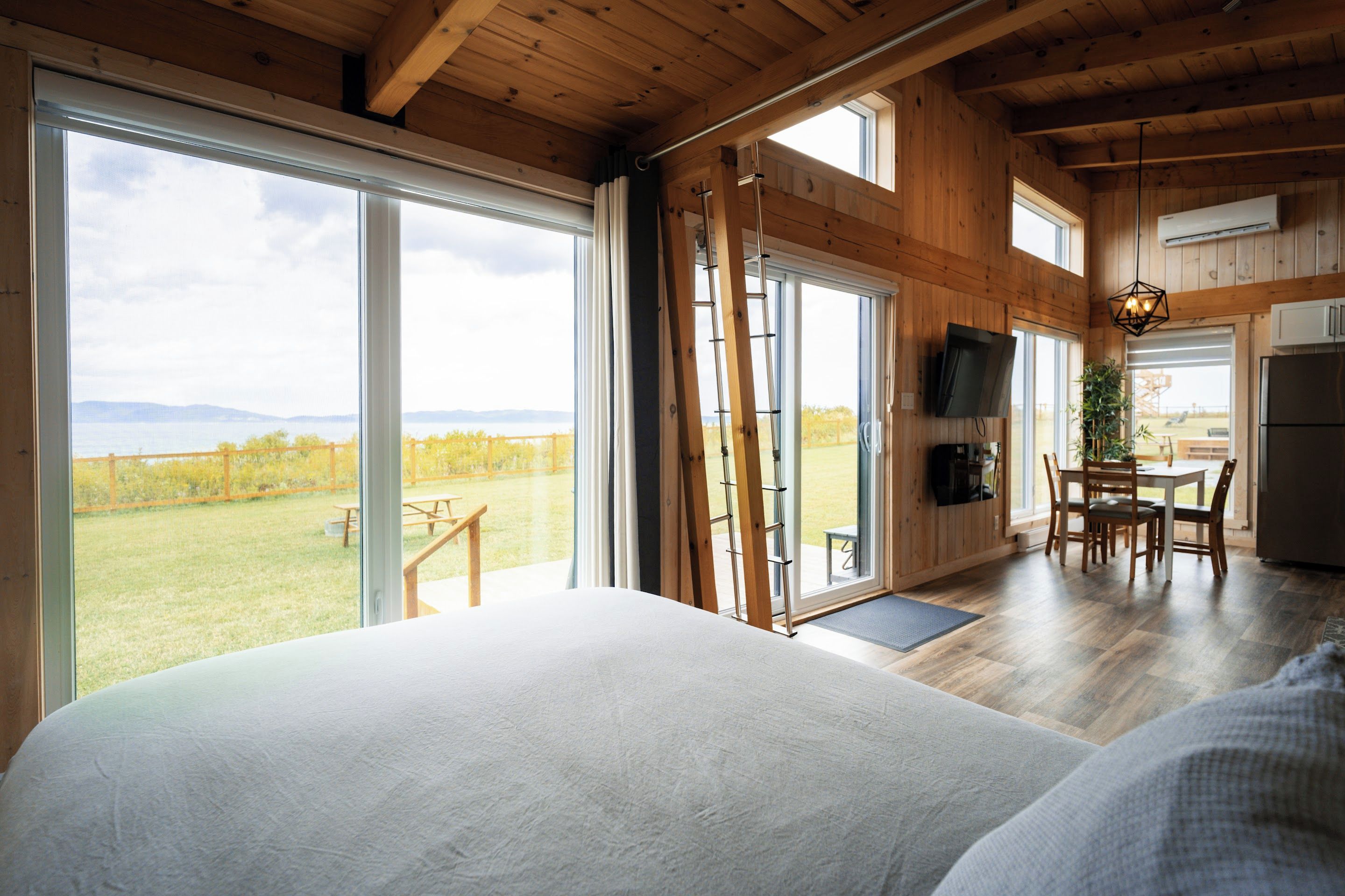 A spacious bedroom with a large bed, a wooden ceiling, and a view of a grassy field through a sliding glass door.