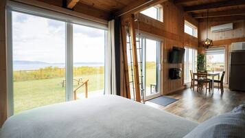 A spacious bedroom with a large bed, a wooden ceiling, and a view of a grassy field through a sliding glass door.