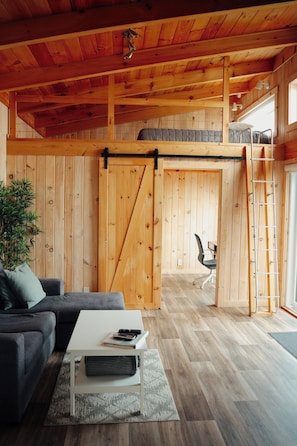 A cozy living room with a sofa, coffee table, and a sliding barn door leading to a loft area with a ladder. - Chalets Nautika (Gaspé)