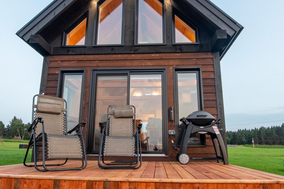 Una cabaña moderna de madera con terraza, dos sillas y una parrilla.