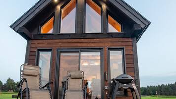 Une cabane en bois moderne dotée d’une terrasse, de deux chaises et d’un barbecue.