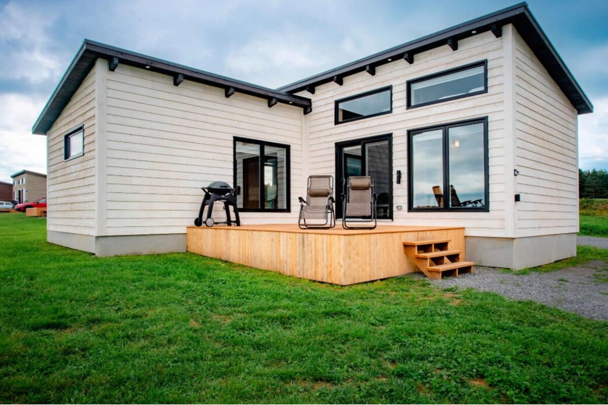 Una casa moderna de dos pisos con una terraza de madera, ventanas grandes y una zona de estar al aire libre cubierta.