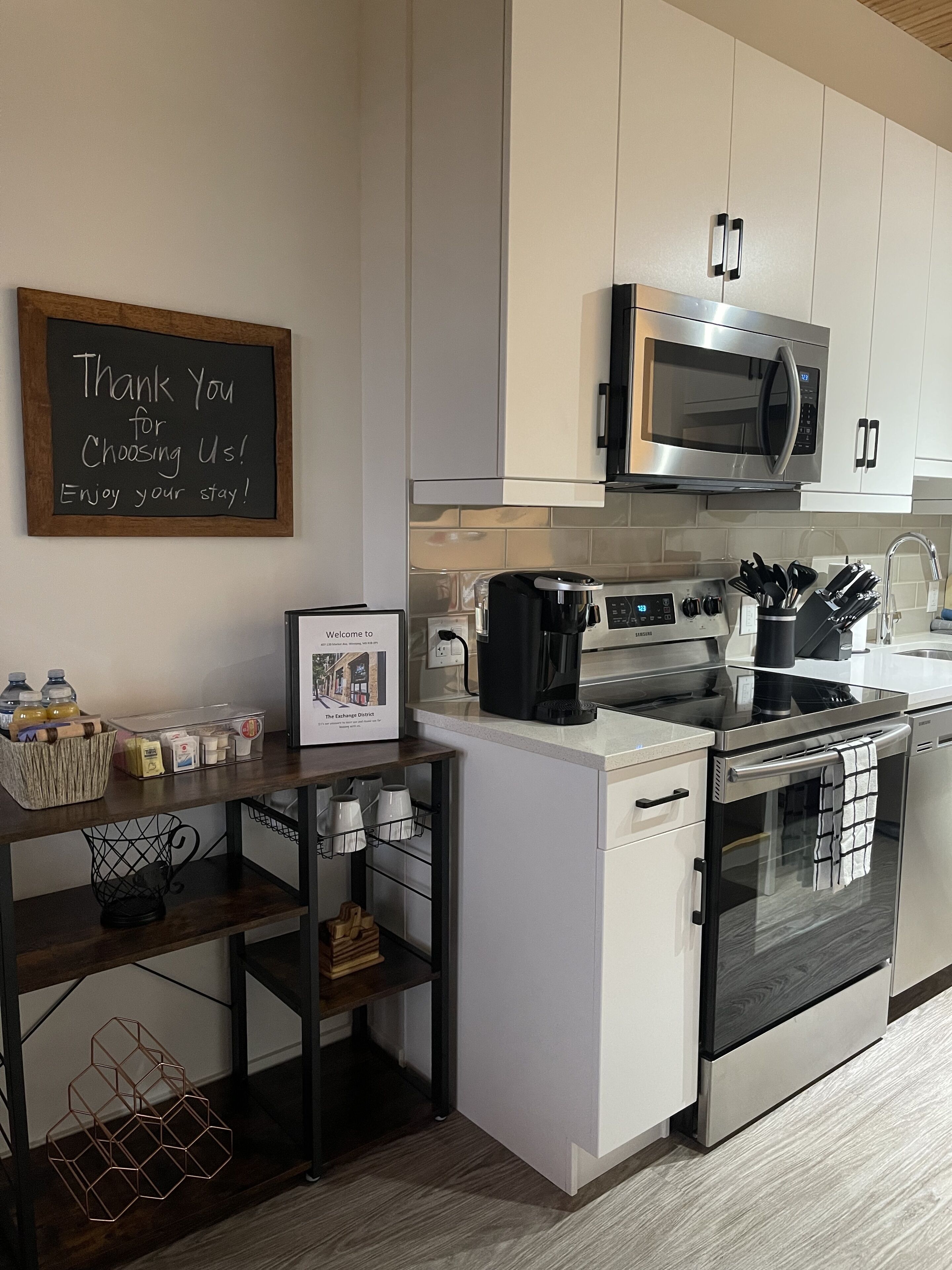 Una cocina moderna con armarios blancos, un microondas de acero inoxidable, una cafetera y un cartel de bienvenida enmarcado.