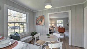 Een eetgedeelte met een ronde tafel, witte stoelen en een bank. Er is een raam met uitzicht naar buiten, een schilderij aan de muur en een plafondlamp.
