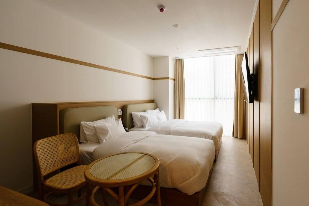 Habitación de hotel con dos camas, una mesita redonda pequeña, una silla de mimbre y un televisor colgado en la pared.
