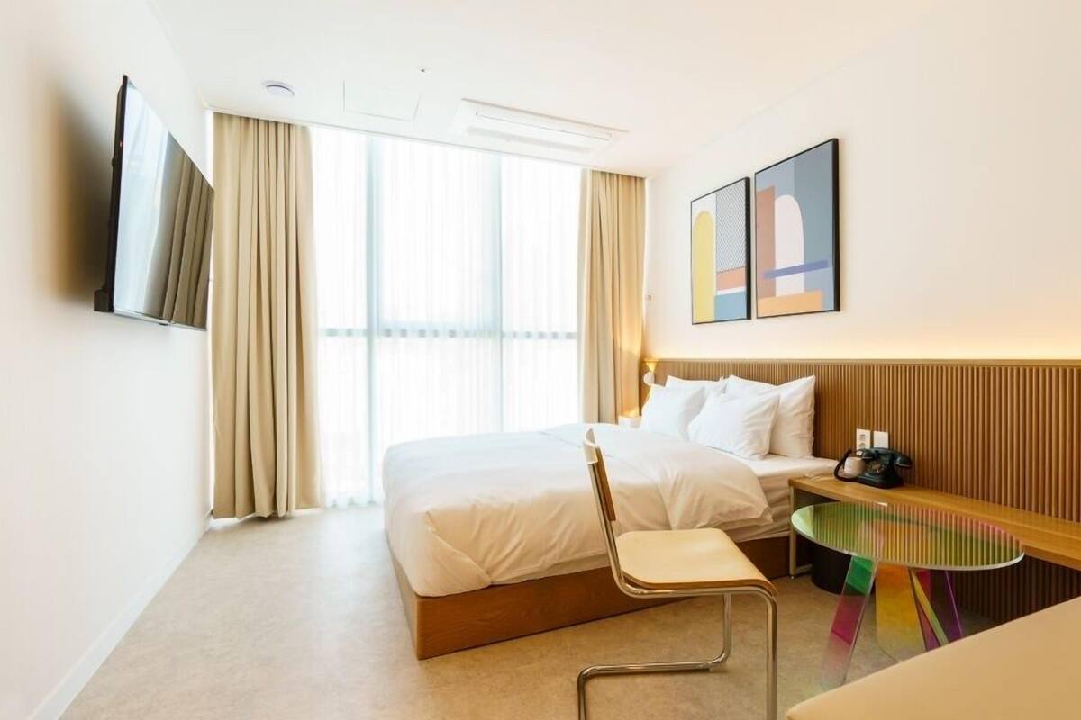 Habitación de hotel con una cama, una silla, una mesita, un televisor y una ventana con cortinas.