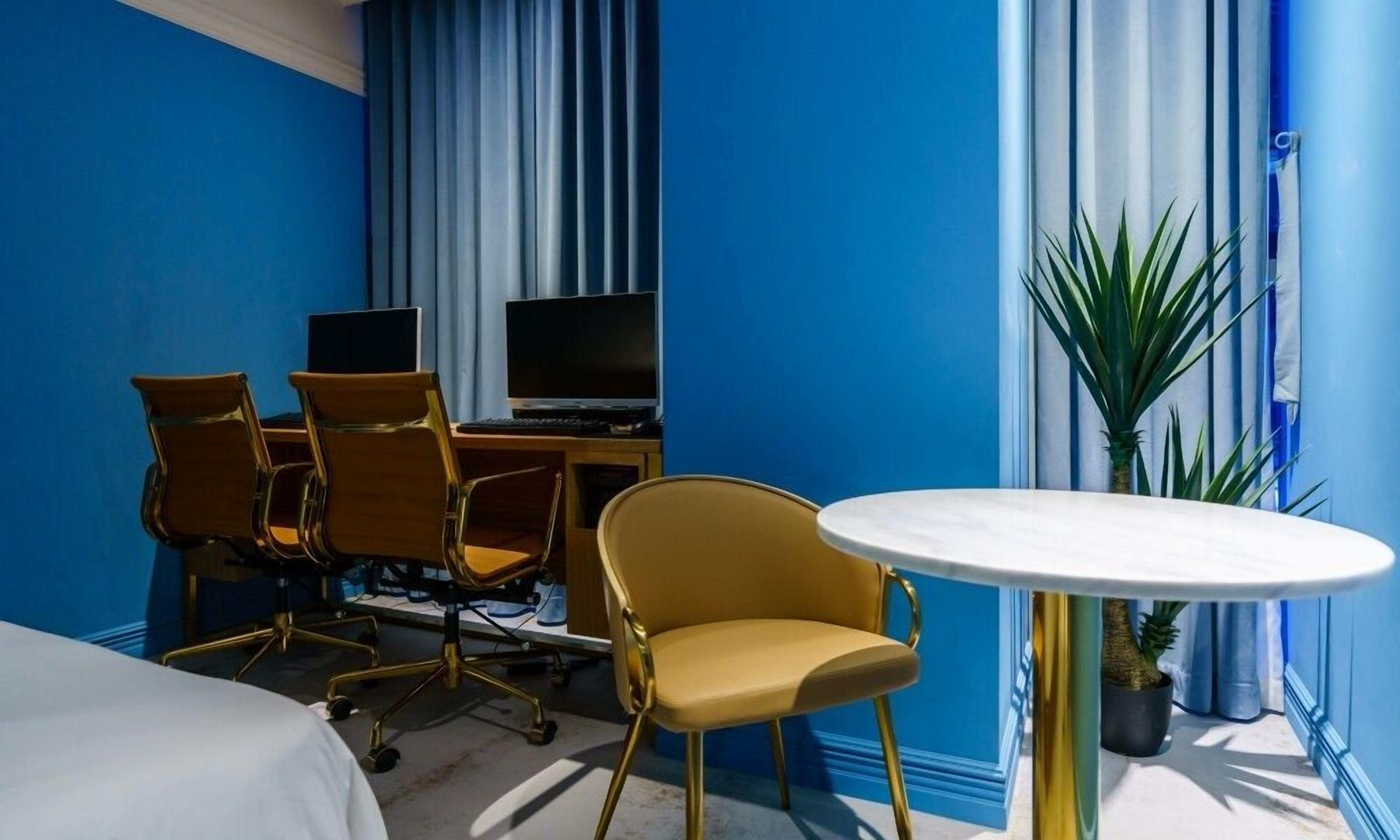 Habitación de hotel con pared azul, una mesa redonda blanca, dos sillas amarillas, un escritorio con ordenador y televisor, y una planta en maceta.