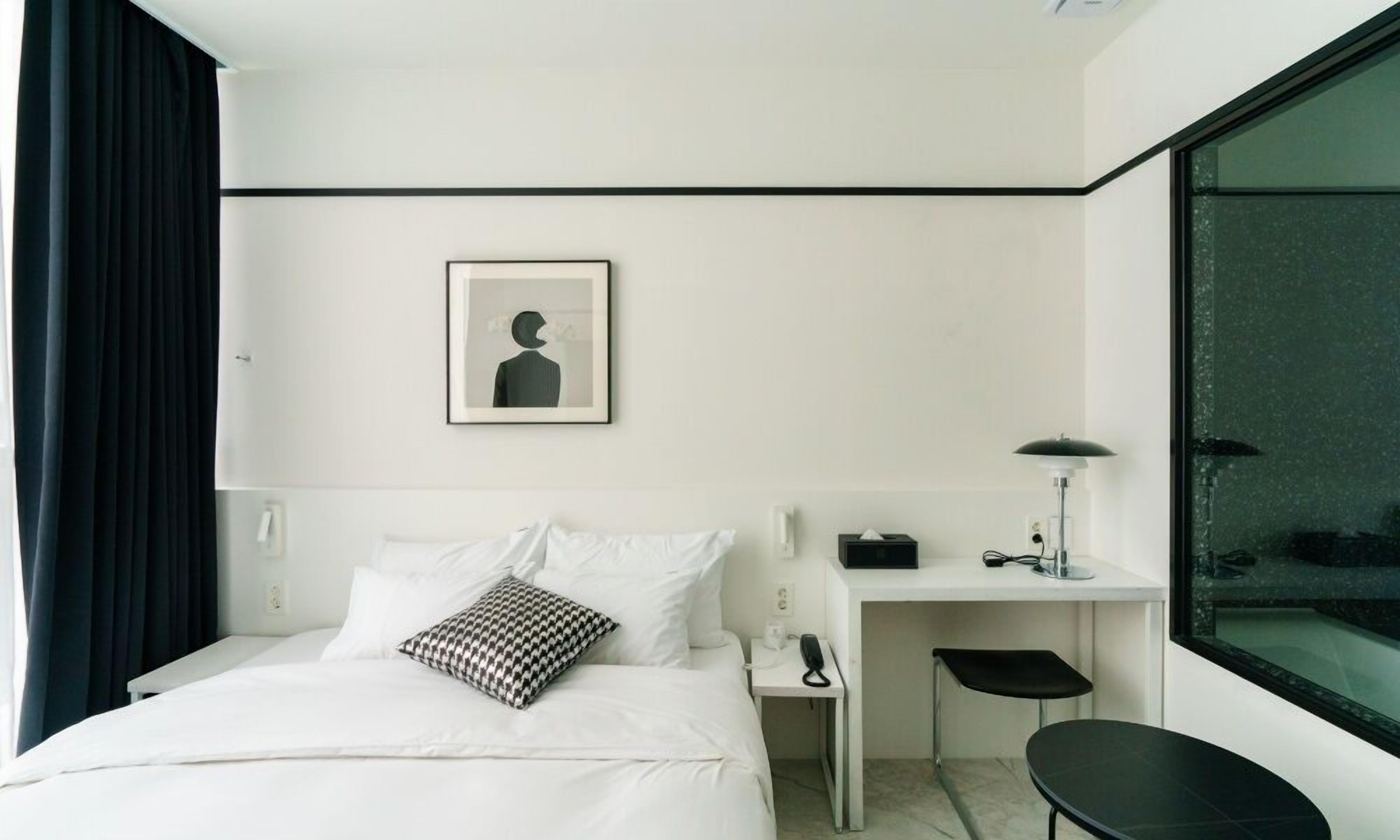 Una habitación de hotel moderna con una cama bien tendida, un escritorio con lámpara, una silla y un ventanal amplio.