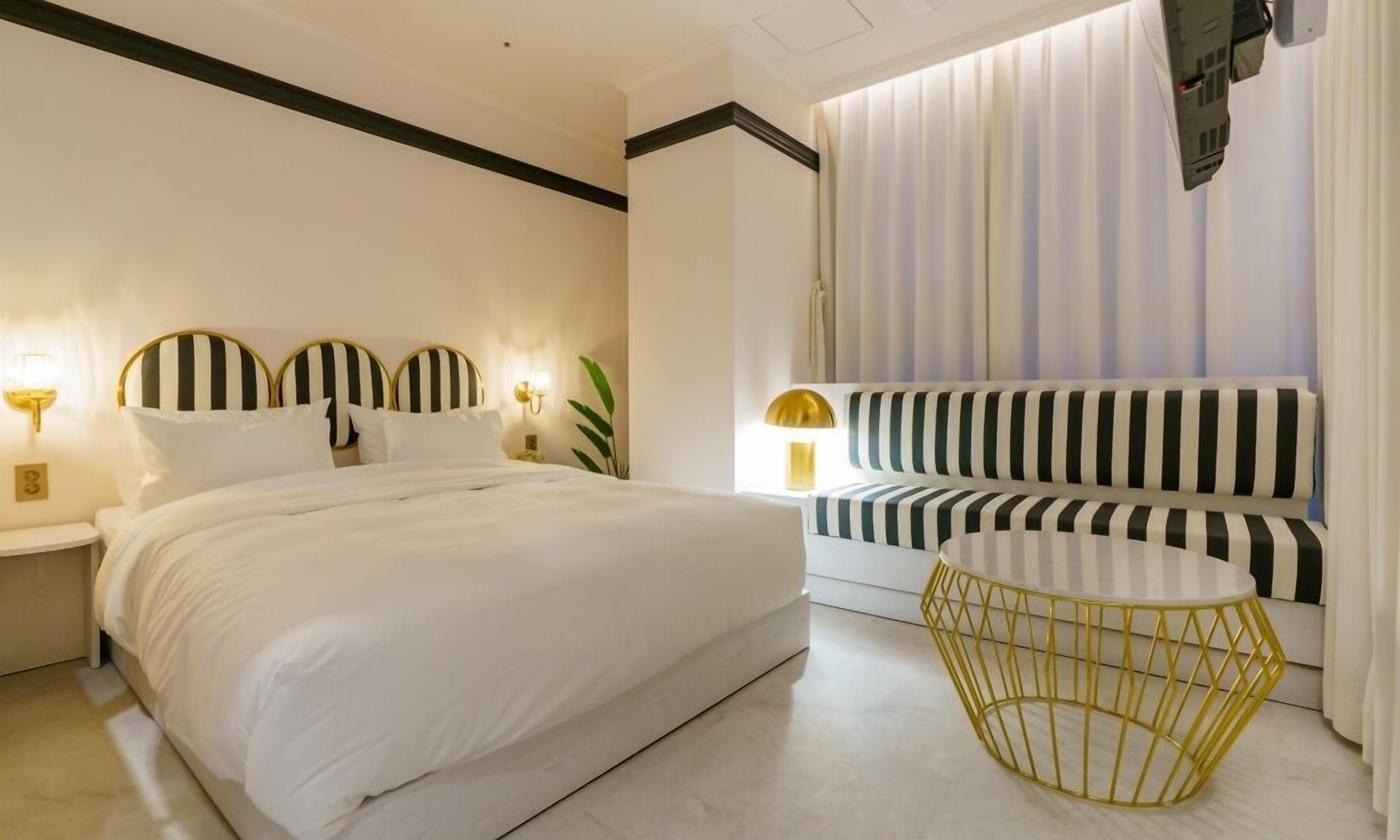 Ein modernes Hotelzimmer mit einem großen Bett, einer Couch, einem runden Tisch mit goldener Rahmung und einem an der Wand befestigten Fernseher.