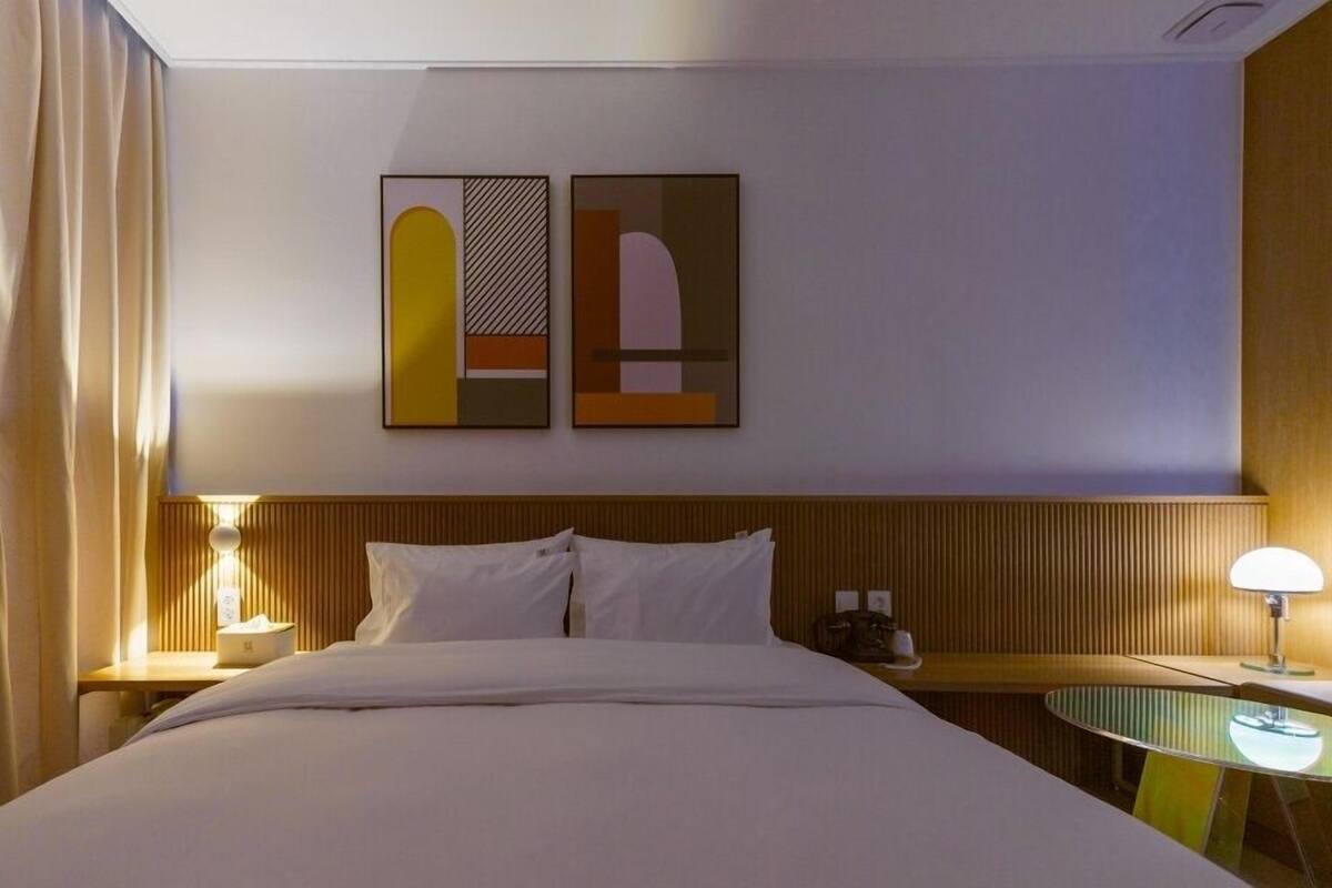 Una habitación de hotel con una cama, dos mesitas de noche, una lámpara de pared y una obra de arte abstracta.