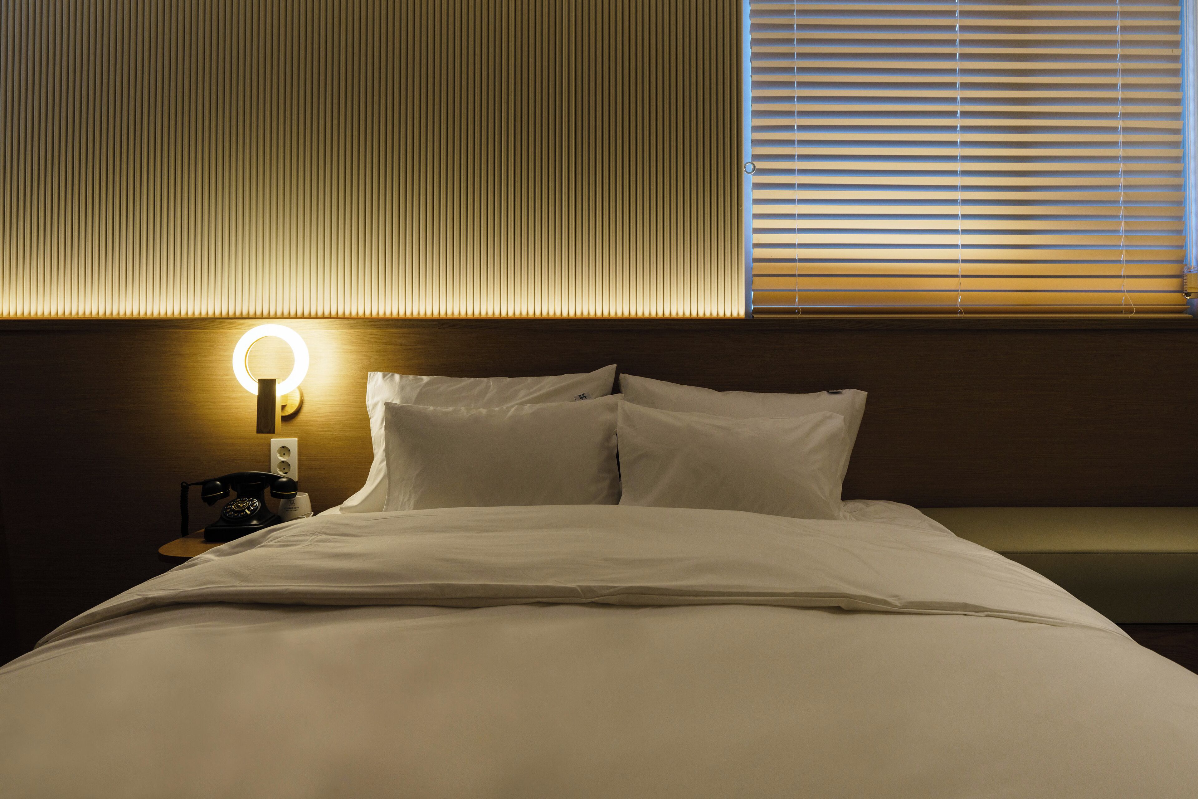 Una cama bien tendida con sábanas blancas, una lámpara de noche y un teléfono sobre una mesita de madera.