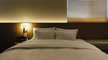 Una cama bien tendida con sábanas blancas, una lámpara de noche y un teléfono sobre una mesita de madera.