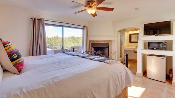 A bedroom with a large bed, a ceiling fan, a fireplace, a small refrigerator, and a view of the outdoors. - 'La Paloma' - Full Circle Ranch Studio w/ Views! (Cave Creek)
