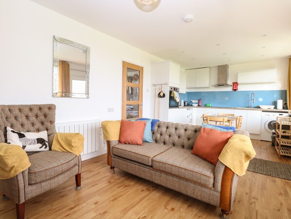 A living room with a sofa and armchairs, a kitchen in the background, and a mirror on the wall.
