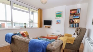 A cozy living room with a sofa, armchair, and a small table. There is a TV mounted on the wall, a bookshelf with books, and a window with curtains. - 31 Seaward Crest (Norwich)