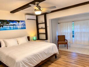 A bedroom with a bed, a ceiling fan, an air conditioning unit, a wall-mounted shelf, a chair, and a view through a window.