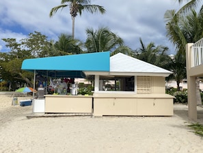 A beachside food kiosk with a blue canopy and beige exterior, surrounded by palm trees and a sandy beach. - VIEWS & MORE VIEWS! Cowpet Bay East, water front condo, steps from beach. (St Thomas)