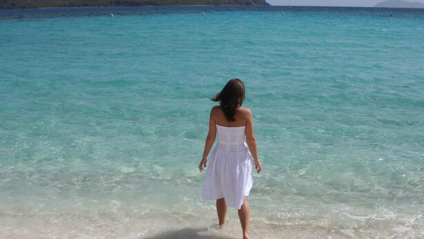 A person in a white dress walking on a sandy beach with clear turquoise water and a distant mountain. - VIEWS & MORE VIEWS!  Cowpet Bay East, water front condo, steps from beach.  (St Thomas)