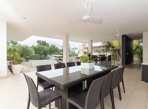 A spacious outdoor dining area with a glass-top table and wicker chairs, surrounded by potted plants and a view of a swimming pool.