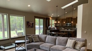 A modern living room with a sectional sofa, a coffee table, and a view of the kitchen and backyard through large windows. - Explore Natures Beauty and Stay at Home in the Heart of Oregon's Wonderland (Bend)