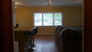 Ein kompakter Wohnbereich mit Küchenzeile, einem Essbereich mit zwei Barhocker, einer Couch und einem großen Fenster mit Blick ins Freie.