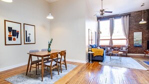 Un vaste espace de vie comprenant une table à manger, des chaises et un canapé. Il y a deux fenêtres avec des rideaux, un ventilateur de plafond et un grand tableau accroché au mur.