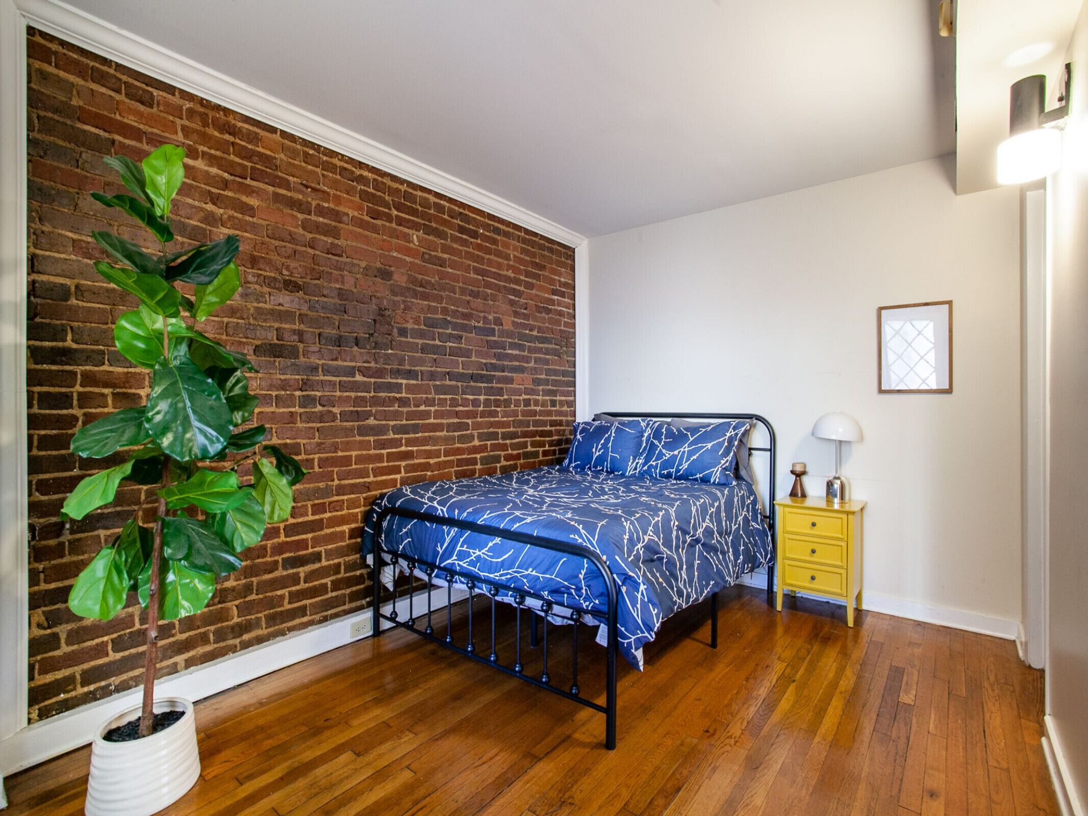 A bedroom with a brick wall, a bed with a blue patterned bedspread, a wooden floor, a yellow nightstand, and a potted plant.