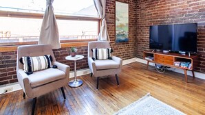Un salon avec deux fauteuils, une petite table ronde, un téléviseur à écran plat posé sur un meuble en bois et un mur en briques.