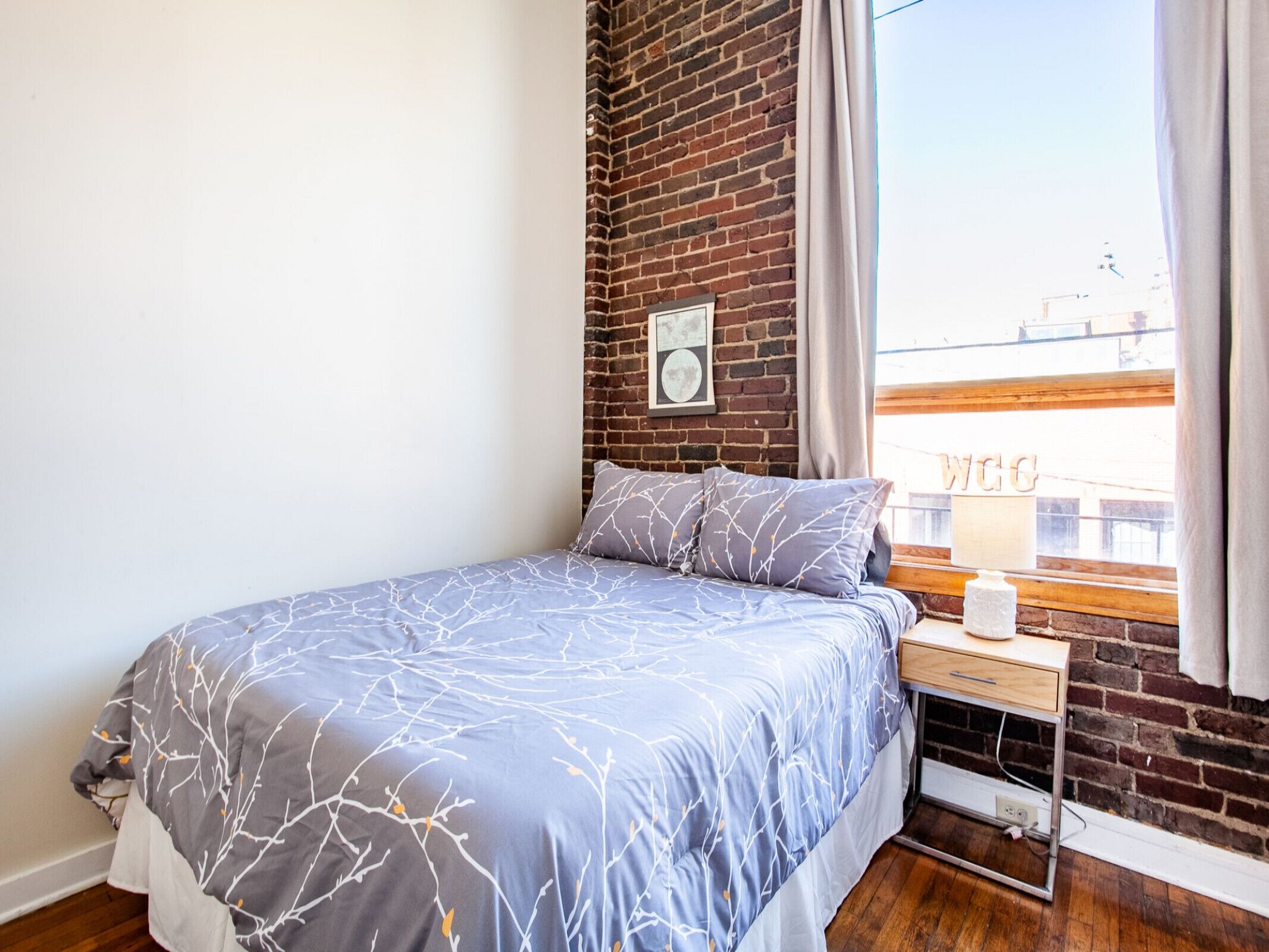 Un dormitorio con cama, mesita de noche, ventana con vista a edificios y pared de ladrillo.