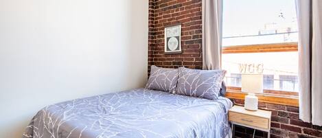 Un dormitorio con cama, mesita de noche, ventana con vista a edificios y pared de ladrillo.