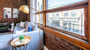 A cozy living room with a brick wall, a large window with a view of buildings, a modern sofa, and a floor lamp. - Modern Old City Loft - Downtown Knoxville (Knoxville)