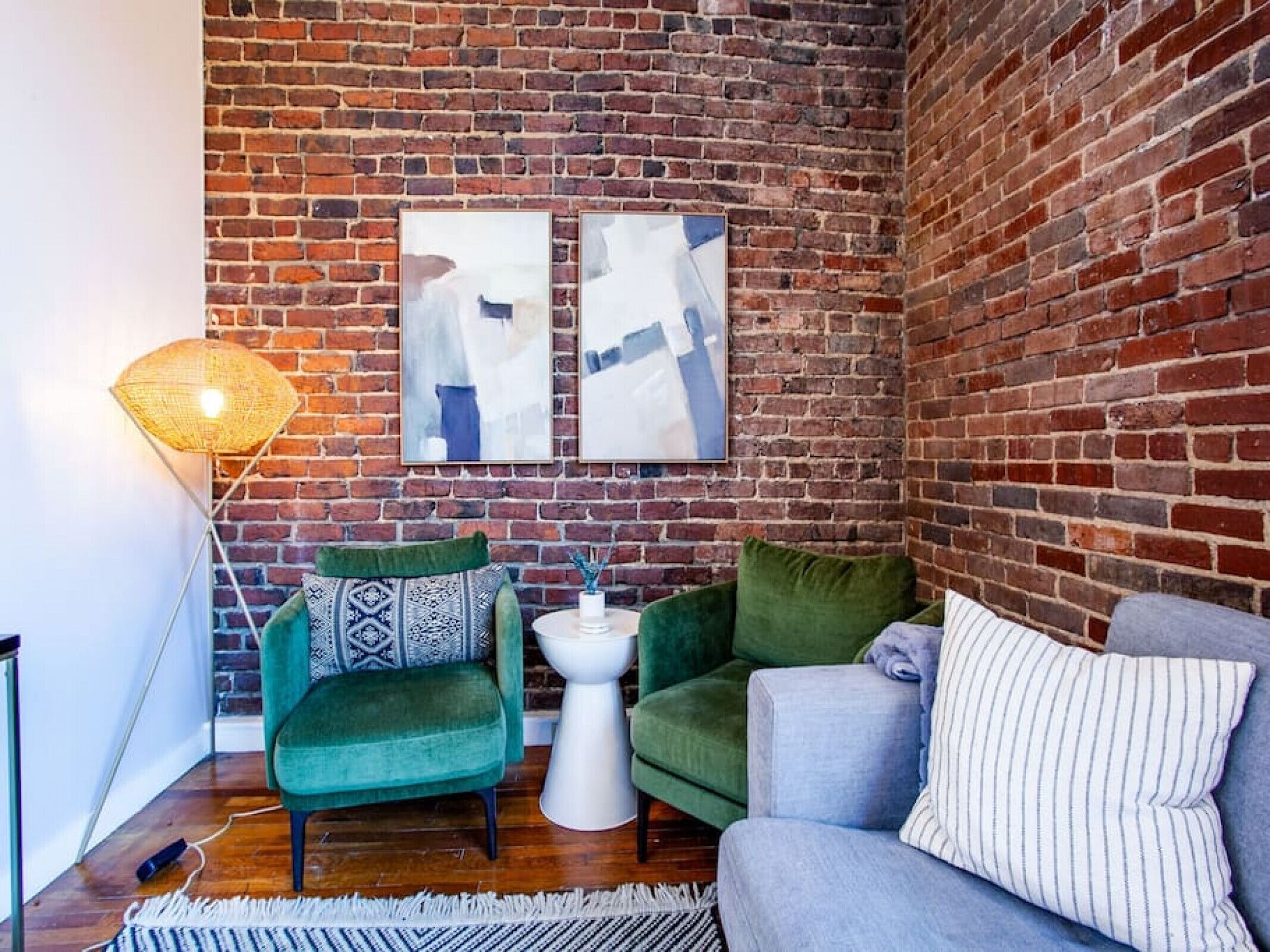 A cozy corner with a brick wall, two green armchairs, a grey sofa, and a white side table with a vase.