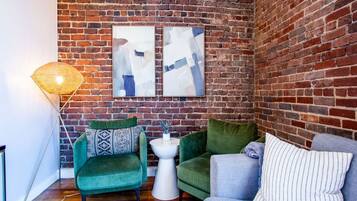 A cozy corner with a brick wall, two green armchairs, a grey sofa, and a white side table with a vase.
