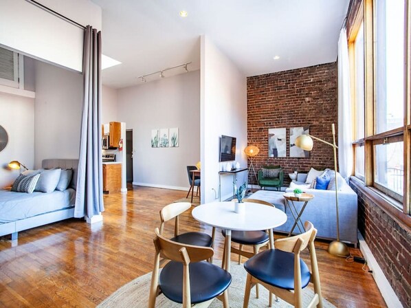 A modern living room with a sofa, a dining area with chairs, a kitchen in the background, and a brick wall.