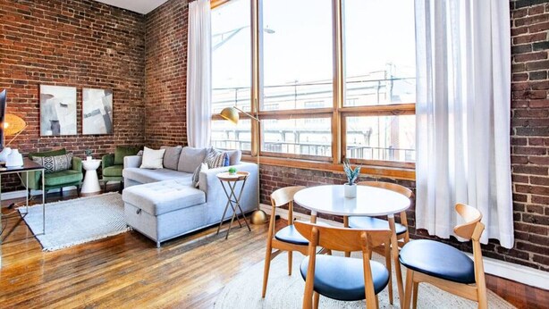 A modern living room with a brick wall, a grey sofa, a round table with chairs, and a large window with curtains. - Modern Old City Loft - Downtown Knoxville (Knoxville)