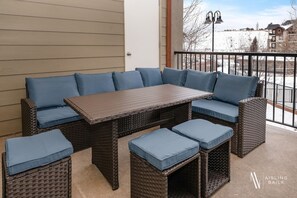 Ein Sitzgruppe aus Rattangeflecht mit blauen Kissen, einem Holztisch und passenden Hocker auf einem Balkon mit Blick auf eine verschneite Landschaft und Gebäude.