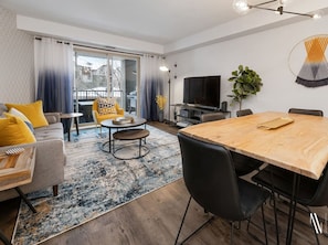 Un salon moderne avec une table à manger en bois, des chaises noires, un canapé gris avec des coussins jaunes, une table basse et un téléviseur.