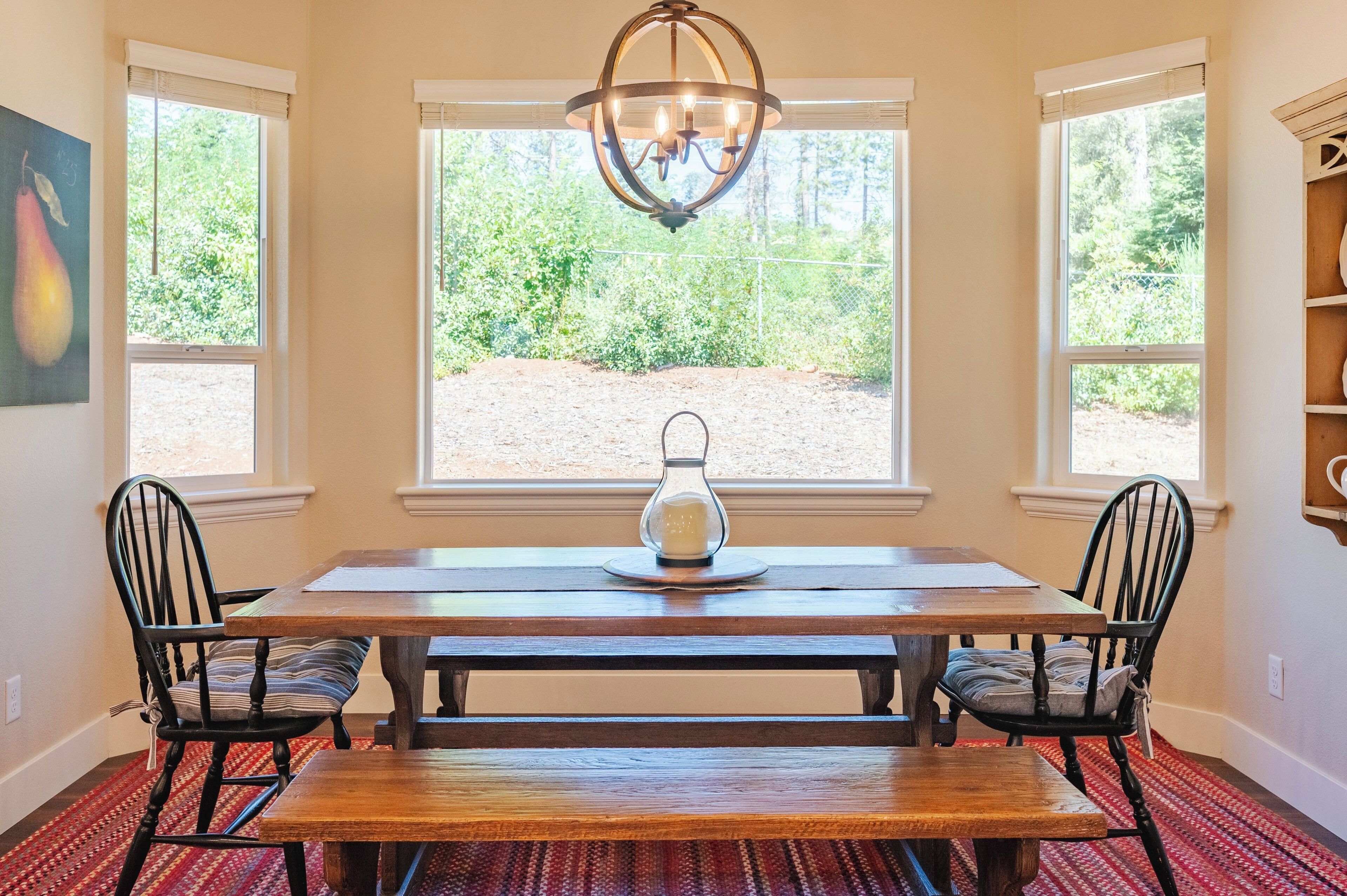 A dining area with a wooden table, two chairs, a chandelier, and a view of greenery through large windows.