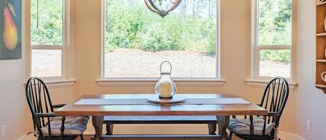 A dining area with a wooden table, two chairs, a chandelier, and a view of greenery through large windows.