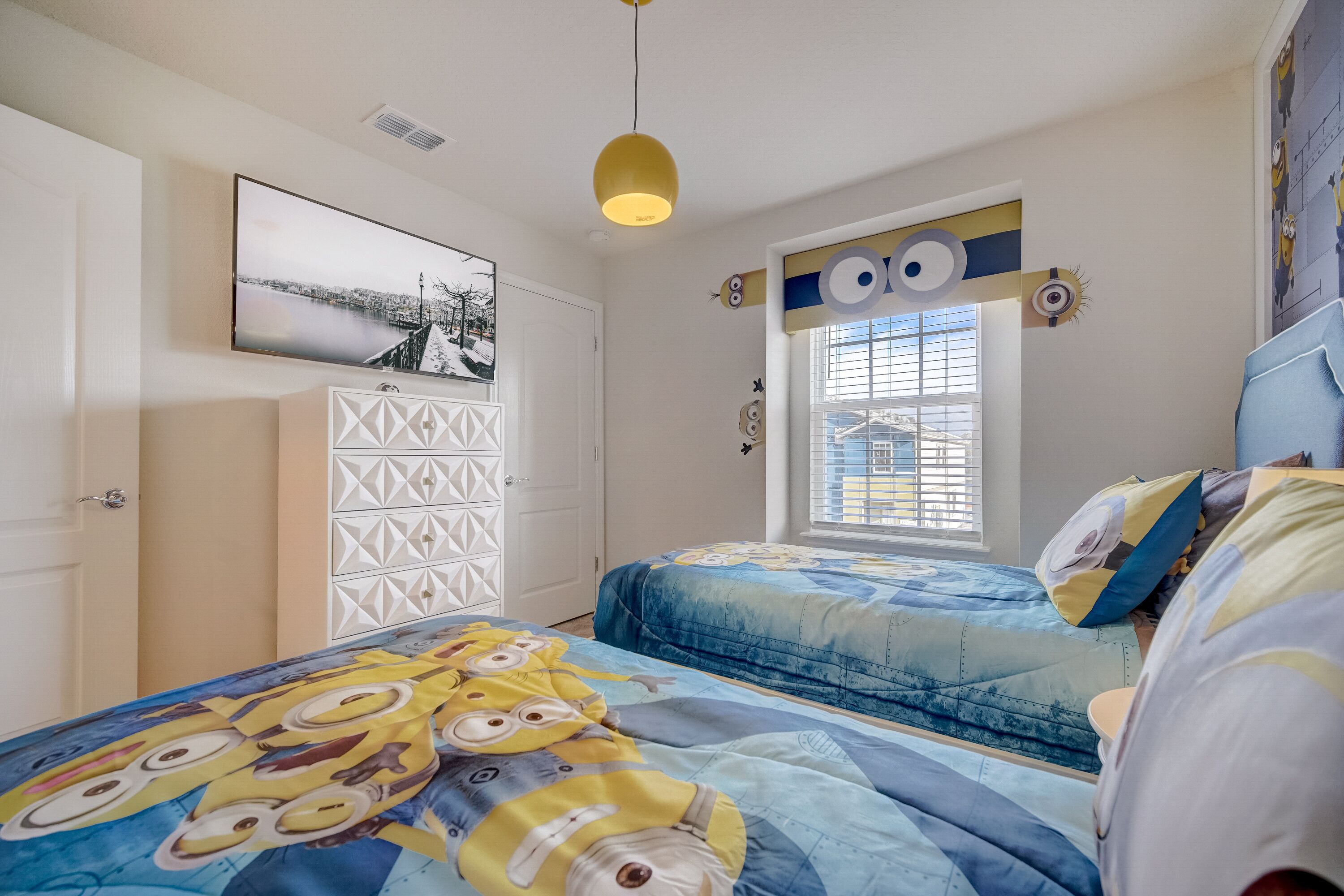 Une chambre avec une couvre-lit aux motifs des personnages de « Moi, moche et méchant », une commode blanche, un tableau encadré au mur et une fenêtre avec des stores.