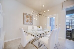 A modern dining area with a glass table, white chairs, a chandelier, and a large abstract painting on the wall.