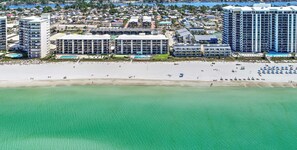 An aerial view of a coastal hotel area with multiple buildings, a sandy beach, and a clear blue sea.
