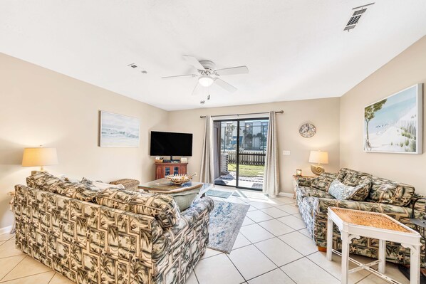 A living room with two patterned sofas, a flat-screen TV, ceiling fan, and a large window with curtains.