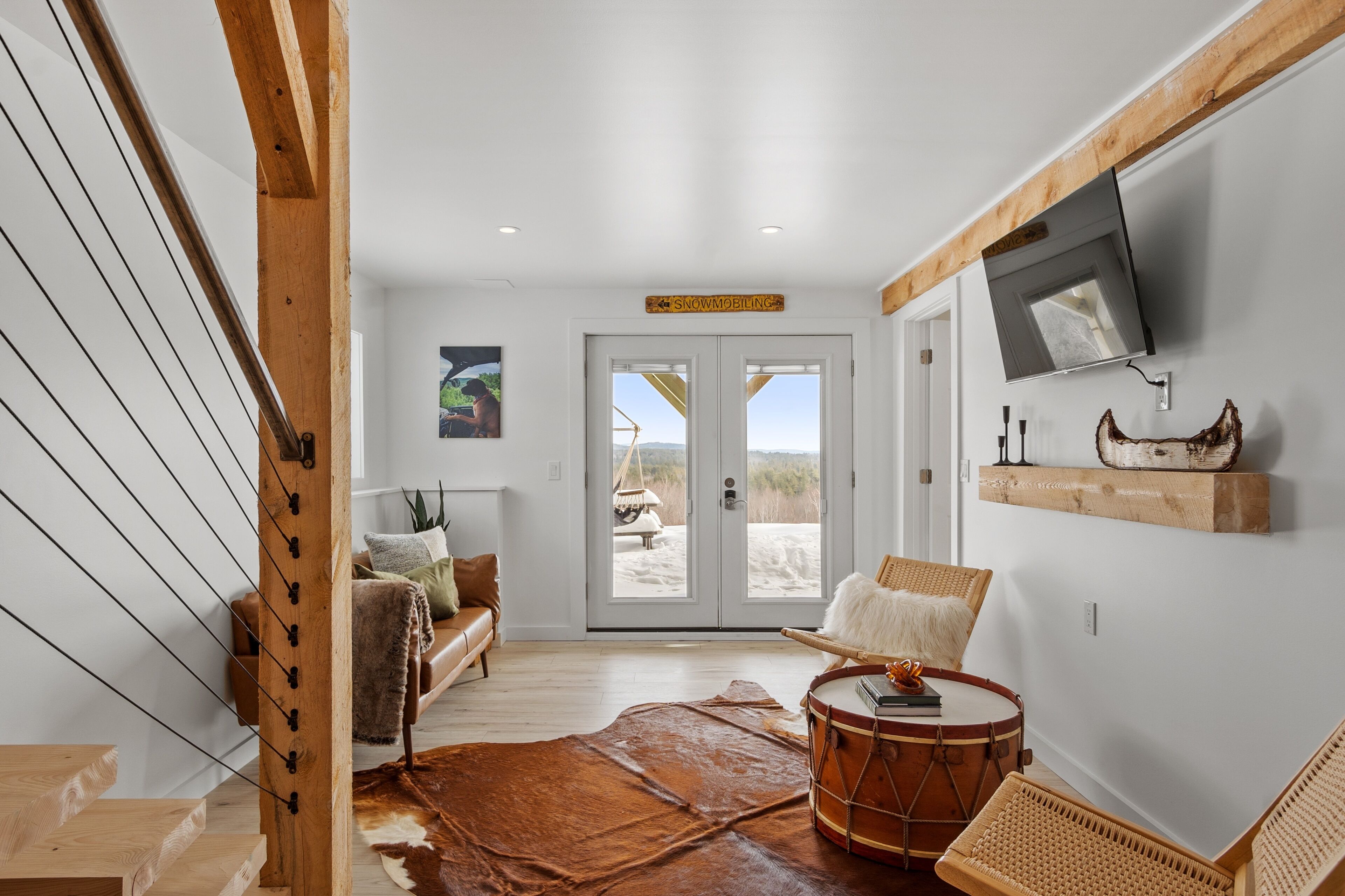 A spacious living room with a large window, a wooden staircase, a television mounted on the wall, and a drum.