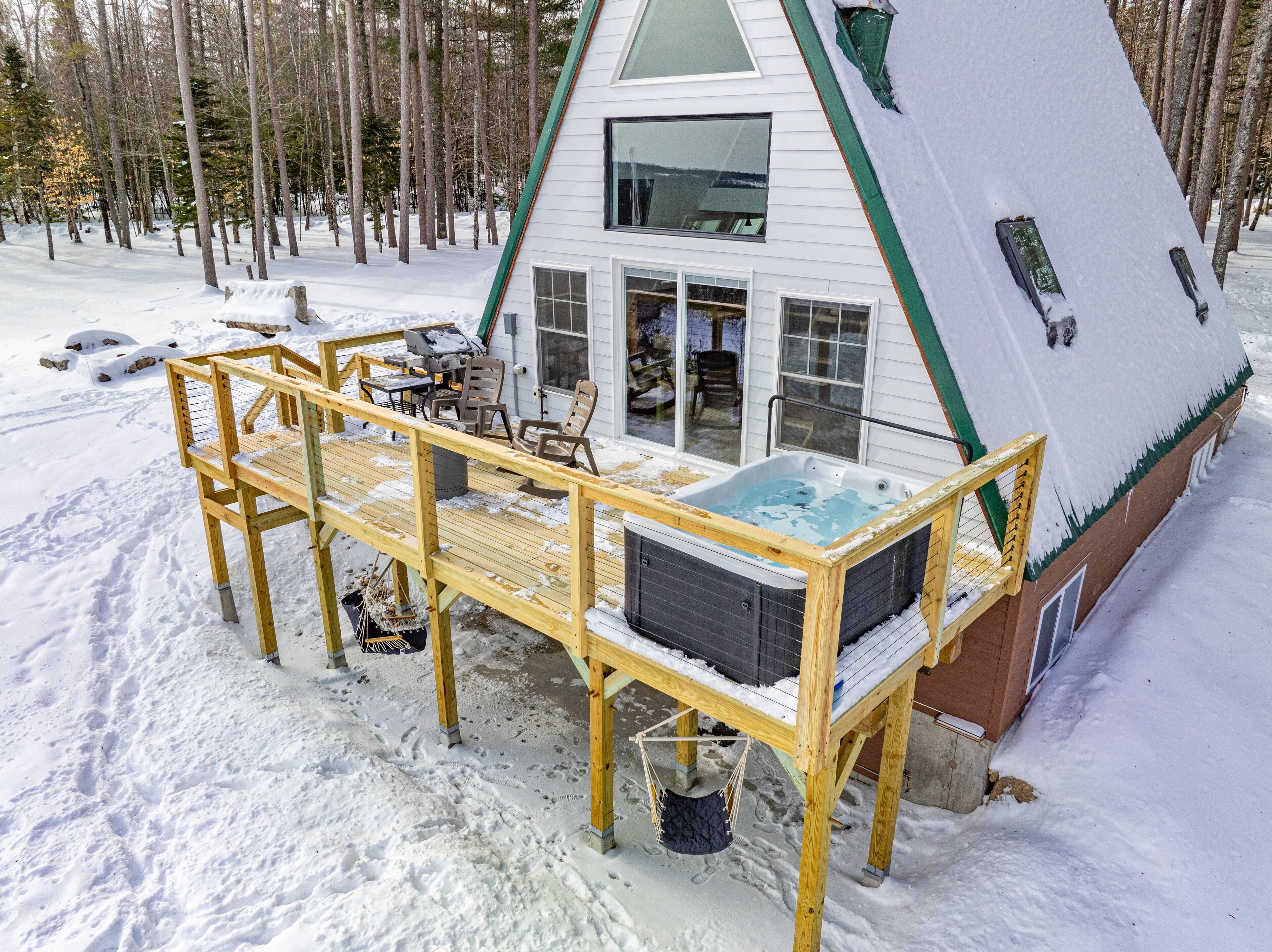 Une terrasse en bois avec un jacuzzi et un espace salon, une grande fenêtre et une vue sur une forêt enneigée.