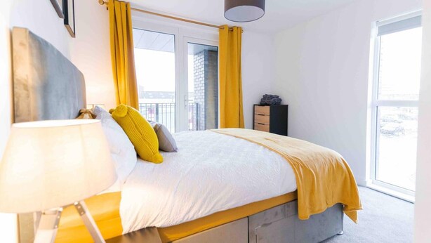 A modern bedroom with a bed, a bedside table, a lamp, and a window with curtains. - Modern Apartment in Salford, Manchester, UK (Salford)