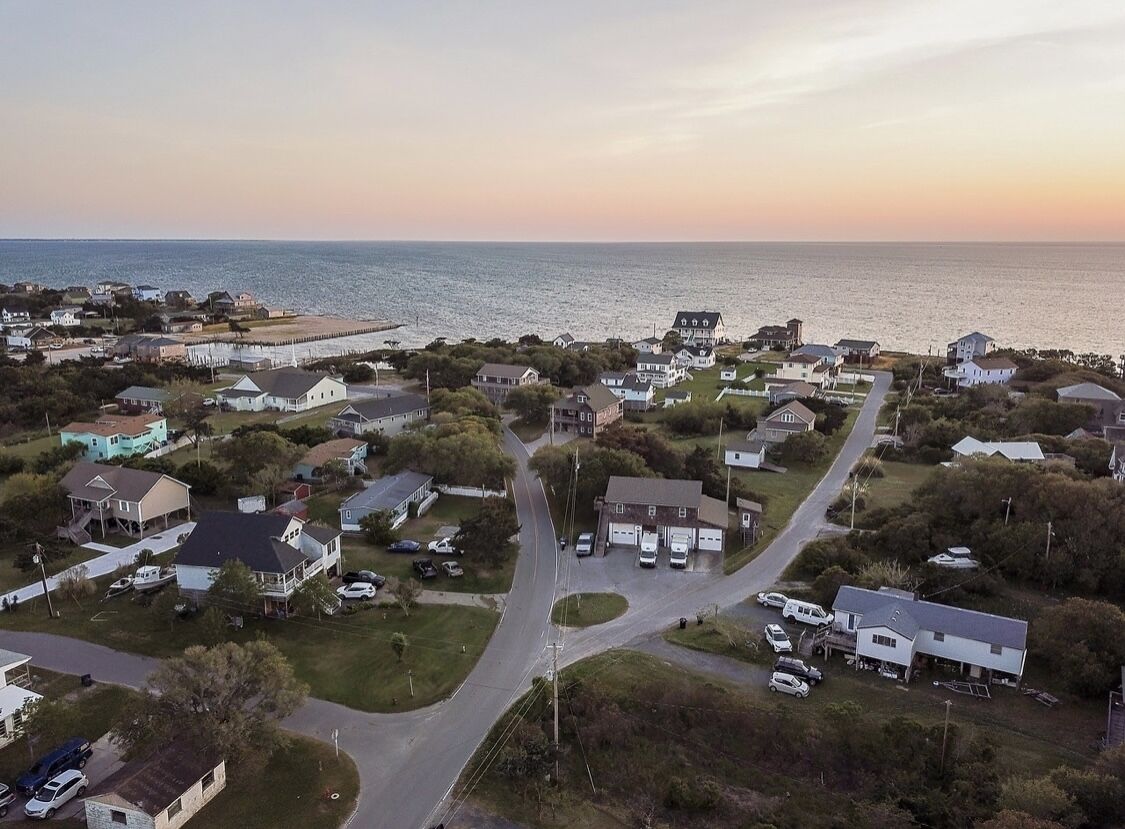 An aerial view of a coastal residential area with houses, roads, and a pier extending into the water.