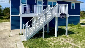 Una casa de playa de dos pisos con una escalera blanca y un letrero que da la bienvenida a visitantes a The Little Gem Beach House en Tops Harbor.